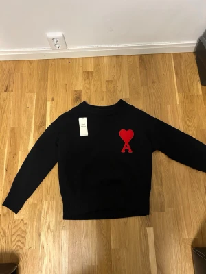 Svart stickad tröja från Ami Paris - Svart stickad tröja från Ami Paris med röd hjärtlogga och bokstaven A på bröstet. Tröjan har rund hals, ribbade muddar och är långärmad. Perfekt för dig som gillar stilrena plagg med en twist.