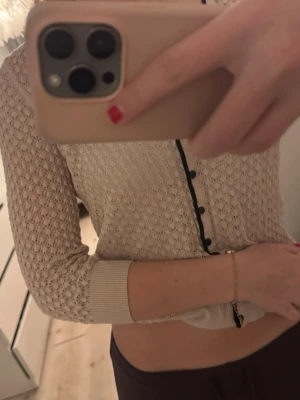 kofta med svarta knappar - Säljer en beige/ljusrosa kofta med snyggt hålmönster och svarta knappar framtill. Koftan har v-ringning med svart kant och ribbade muddar vid ärmslut och nederkant. Perfekt att slänga över ett linne för en chill look.