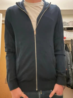 Mörkblå hoodie med dragkedja - Snygg mörkblå hoodie med hel dragkedja framtill och huva. Tröjan har långa ärmar och ribbade muddar vid ärmslut och nederkant. Perfekt för chill dagar och enkel att matcha med jeans eller joggers. Materialet känns mjukt och bekvämt mot huden.