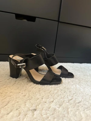 Svarta klack-sandaler Steven New York - Säljer ett par svarta sandaler från Steven New York i storlek 36. Skorna har breda remmar, öppen tå och hög klack. Tillverkade i skinn med spänne vid ankeln. Perfekta för dig som gillar stilrena och klassiska vibes.