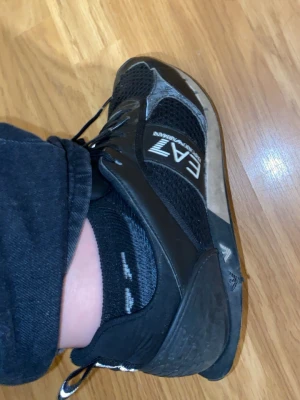 Svarta sneakers från Emporio Armani - Säljer ett par svarta sneakers från Emporio Armani EA7 med meshpaneler och snygga logodetaljer på sidan. Skorna har snörning och en platt sula, perfekt för dig som gillar stilrena och sportiga skor. Materialet är en mix av syntet och mesh.