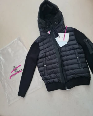 Svart jacka-Moncler -                              Storlek : S                                                        Svart pufferjacka från Moncler med stickade ärmar och huva. Jackan har quiltad front i dun, dragkedja, ribbade muddar och två fickor med tryckknappar. Moncler-logga på ärmen och snygga detaljer. Perfekt för kyliga dagar och riktigt stilren.