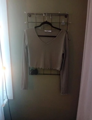 Grön croppad långärmad topp NA-KD - Trendig grön croppad topp från NA-KD med lång ärm och v-ringad hals. Toppen har vågig kant nertill och vid ärmslut, vilket ger en cool och avslappnad look. Perfekt att matcha med höga jeans eller kjol.