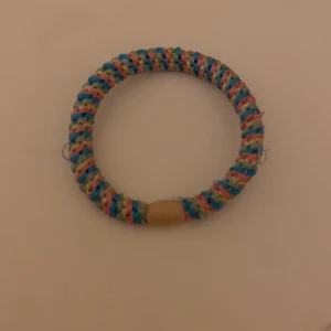 Färgglatt flätat armband - Säljer ett runt armband med flätad design i blått, rosa och beige. Armbandet har en enkel och avslappnad stil, perfekt för att addera lite färg till din look. Det är gjort av syntetmaterial och har en liten beige detalj som avslut.