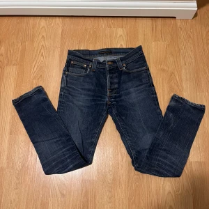 Mörkblå jeans från Nudie Jeans - Snygga mörkblå jeans från Nudie Jeans med klassisk femficksdesign och kontrastsömmar. Modellen har raka ben och subtila slitningar för en cool look. Bakfickorna har Nudie Jeans signaturbroderi och läderpatch i midjan.