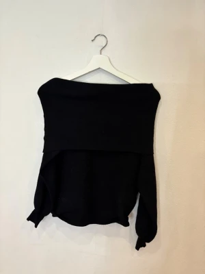 Svart offshoulder stickad tröja - Svart stickad tröja med offshoulder-design och långa ärmar. Tröjan har en bred krage som vilar över axlarna och ger en cool, avslappnad vibe. Perfekt för dig som gillar stilrena och enkla plagg med en twist.