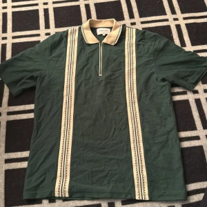 Grön henley från Palmes med krage - Grön henley från Palmes med beige krage och dragkedja framtill. Skjortan har korta ärmar och två beige ränder med mönster längs framsidan. Perfekt för dig som gillar retrovibbar och snygga detaljer.