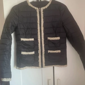 Svart quiltad pufferjacka från Mango - Quiltad svart pufferjacka från Mango med beige volangdetaljer längs kanter, fickor och ärmslut. Jackan har rund hals, dragkedja framtill och fyra fickor med dekorativa beigea kanter. Snygg och stilren modell som ger en cool vibe.