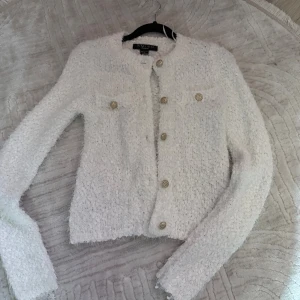 Vit cardigan jacka från Amisu XS - Säljer en fluffig vit cardigan jacka från Amisu i storlek XS. Jackan har snygga guldknappar framtill och på bröstfickorna, rund hals och lång ärm. Materialet är stickat och ger en mysig och trendig look, perfekt för att lyfta din outfit.