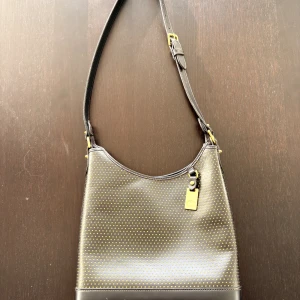 Svart axelväska från Dooney & Bourke - Snygg svart axelväska i skinn från Dooney & Bourke med guldiga detaljer och små prickar i mönstret. Väskan har justerbar axelrem, dragkedja upptill och metallfötter undertill. Perfekt för dig som vill ha en stilren och unik väska med plats för det viktigaste.