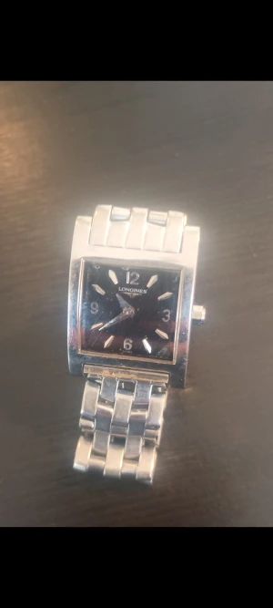 Longines fyrkantig silverklocka - Säljer en stilren Longines Dolce Vita klocka med fyrkantigt ur och svart urtavla. Armbandet är i silverfärgat metall med klassisk länkdesign. Klockan har tydliga siffror och markeringar på urtavlan, och en smidig låsmekanism. Perfekt för dig som gillar tidlös design. Länken har släppt från låset en kvick fix för en kunnig person. Det vill säga 50 spänn hos en klick handlare.