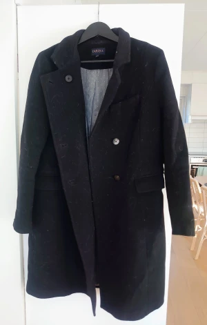Svart ullkappa från Caroll, stl 42 - Svart kappa från Caroll med klassisk krage och knäppning framtill. Tillverkad i ull med snyggt randigt och prickigt foder i grått. Kappan har två fickor med lock och långärmade ärmar. Perfekt för dig som gillar stilrena och tidlösa plagg.