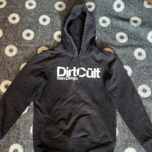 Svart hoodie DirtCult  - Svart hoodie från DirtCult med stor vit logga på bröstet och texten San Diego. Tröjan har huva med snörning och en klassisk magficka. Perfekt för chill dagar och streetwear-stil. Långärmad och i mjukt bomullsmaterial.