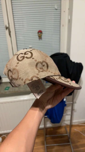 Beige Gucci keps med GG-mönster - Säljer en beige keps från Gucci med det klassiska GG-mönstret i brunt. Kepsen har justerbar rem i mörkbrunt skinn baktill och är tillverkad i slitstark bomull. Perfekt för dig som vill ha en lyxig och snygg accessoar med street-vibe.