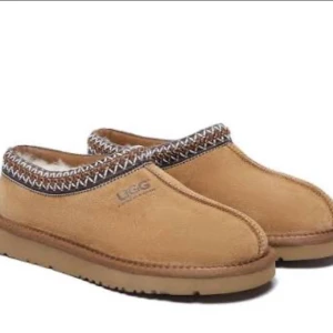 Äkta sheperd UGG beige slip-on boots - Säljer ett par beige slip-on boots från UGG med rund tå och platt sula. Skorna har ett snyggt mönstrat band runt öppningen och är fodrade med mjuk päls för extra komfort. Perfekta för kalla dagar och har en klassisk UGG-look.