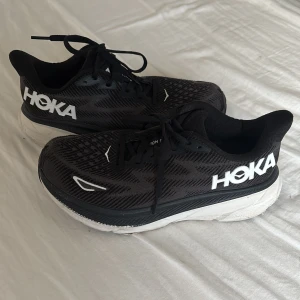 Svarta Hoka Clifton 9 sneakers - Säljer ett par svarta Hoka Clifton 9 sneakers med vit och svart tjock sula. Skorna har snörning och är tillverkade i syntetmaterial med ventilerande mesh-överdel. Hoka-loggan syns tydligt på sidan i vitt. Perfekta för dig som gillar att springa eller träna.