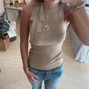 Beige ribbad linnetopp med knyt - Säljer en beige ribbad linnetopp med snygg knyt-detalj vid axeln. Toppen är ärmlös och har en tight passform som framhäver figuren. Perfekt för dig som gillar stilrena och trendiga plagg med det lilla extra.