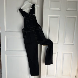 Svarta denim hängselbyxor från Monki - Svarta hängselbyxor i denim från Monki med kontrasterande vita sömmar. Klassisk modell med stor bröstficka, knappar i sidorna och flera fickor bak. Justerbara axelband och avslappnad passform. Perfekta för en chill och trendig look.