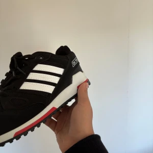 Adidas ZX 750 svarta sneakers - Adidas ZX 750 sneakers i svart med vita ränder och röda detaljer på sulan. Skon har snörning, platt sula med grovt mönster och rund tå. Materialet är en mix av syntet och mocka, med meshpartier för extra ventilation. Ikonisk Adidas-logga på hälen och sidan.