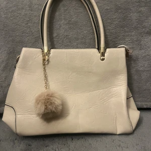 Beige handväska med fluffig detalj - Stilren beige handväska med guldfärgade detaljer och en fluffig beige boll som hänger i kedja. Väskan har flera fack med dragkedjor och svarta kanter på handtagen. 