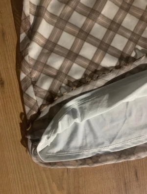 Brun och vit rutig minikjol - Supersöt kortkjol med rutigt mönster i brunt och vitt. Kjolen har en liten rosett framtill och volangkant längst ner för extra detalj. Tillverkad i mjukt syntetmaterial med vit insida. Perfekt för dig som gillar preppy och trendiga vibes.