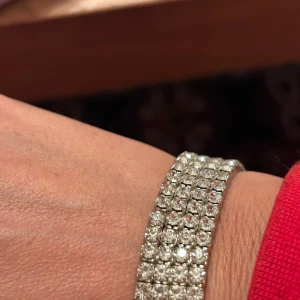 Bred glittrigt armband i silver - Säljer ett brett armband med flera rader gnistrande stenar som ger en riktigt glittrig look. Armbandet är i silverfärg och har en lyxig känsla med sina tätt sittande, fyrkantiga stenar. Perfekt för dig som vill sticka ut och addera extra bling till din outfit.