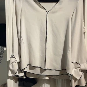 Vit blus med svarta kanter och rosetter - Supersöt vit blus med svarta kontrastsömmar och stora rosetter på ärmarna. Blusen har v-ringning och långa ärmar med volangdetaljer. Perfekt för dig som vill sticka ut med en unik och trendig look.