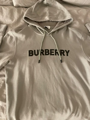 Grå hoodie från Burberry med tryck - Snygg grå hoodie från Burberry med svart logotyptryck på bröstet. Tröjan har huva med snörning och långa ärmar. Tillverkad i mjuk bomull som är skön att ha på sig. 