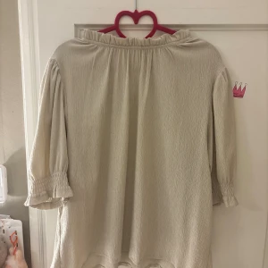 Beige blus  - En snygg beige blus med polyester tyg i storlek L från HOLLY & WHYTE, den är ganska stretchig, någon typ av volang i slutet av armarna och vid halsen med en knapp på baksidan 