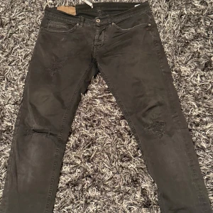Svarta jeans från Dondup, storlek 33 - Svarta jeans från Dondup med slitna detaljer och trasiga partier på benen för en cool, edgy look. Klassisk femficksmodell med knappgylf och raka ben. Jeansen är tillverkade i bomull och har en snygg mörk tvätt. Skriv vid frågor och priset kan diskuteras!