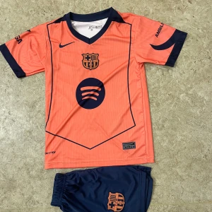 Barça Lamine Yamal fotbollströja orange - Snygg FC Barcelona fotbollströja med Lamine Yamal och nummer 10 på ryggen. Tröjan är orange med mörkblå detaljer, korta ärmar och V-ringad krage. Materialet är lätt och andas, perfekt för träning eller match. Barca-logga och sponsortryck på bröstet.