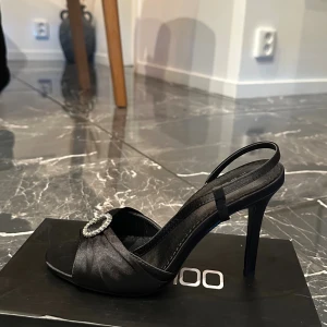 Svarta klack-sandaler från Boohoo - Snygga svarta sandaler med hög klack från Boohoo. Skorna har öppen tå, smal rem runt hälen och en glittrig dekoration framtill. Materialet är satinliknande och ger en lyxig känsla.