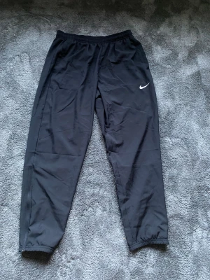 Svarta Nike Dri-FIT träningsbyxor L - Svarta träningsbyxor från Nike med Dri-FIT-teknologi. Byxorna har elastisk midja, smal passform och dragkedja vid benslut. Swoosh-logga på låret. Perfekta för dig som vill ha bekväma och funktionella byxor till träning. Säljer då jag köpte fel storlek och kom på de försent. Använda tre gånger.
