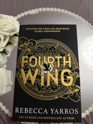 Fourth Wing - En global succé som fångar dig direkt! Fourth Wing av Rebecca Yarros har en snygg, mystisk omslagsdesign med guld och svart, perfekt för dig som älskar episka äventyr och spänning. En bok som snabbt blivit en favorit bland unga vuxna.