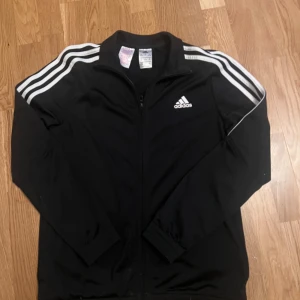 Svart Adidas vindjacka med vita ränder - Svart vindjacka från Adidas med klassiska vita ränder längs axlarna och ärmarna. Jackan har hel dragkedja framtill, ståkrage och Adidas-logga på bröstet. Perfekt för sport eller chill, stilren och enkel att matcha med dina favoritjeans.