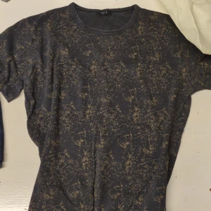 Filippa K - Svart t-shirt med mönster - Cool svart t-shirt från Filippa K i storlek M med unikt abstrakt mönster i beige över hela plagget. T-shirten har rund hals och korta ärmar, tillverkad i mjuk bomull som känns skön mot huden. Perfekt för dig som gillar stilrena detaljer med edge.