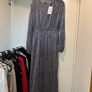 Lång glittrig silverklänning med paljetter - Säljer en maxilång silverfärgad klänning från NAKD, täckt av glittrande paljetter. Klänningen har långa ärmar med lätt puff och markerad midja för en snygg siluett. Perfekt för dig som vill sticka ut med en riktigt glam look!
