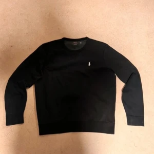 Svart sweatshirt från Polo Ralph Lauren - Svart sweatshirt från Polo Ralph Lauren med klassisk rund hals och lång ärm. Tröjan har en vit broderad logga på bröstet och är tillverkad i mjuk bomull. Perfekt för en chill och stilren look.