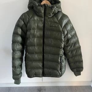 Grön Peak Performance pufferjacka M - Säljer en grön pufferjacka från Peak Performance i storlek M. Jackan har huva, dragkedja framtill och två sidofickor med dragkedja. Den är quiltad och har elastiska muddar vid ärmslut. Perfekt för kalla dagar och har en clean, sportig look.