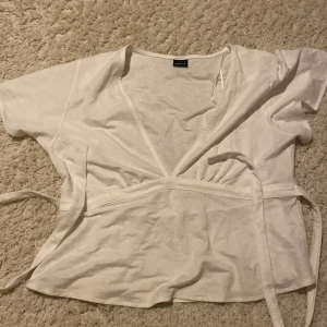 Vit omlottopp med knyt från H&M - Säljer en vit omlottopp från Gina tricot  med korta ärmar och knytband i midjan. Toppen har v-ringad hals och är gjord i mjuk bomull, perfekt för en chill och clean look. Enkel att styla med jeans eller kjol för en fräsch vibe.