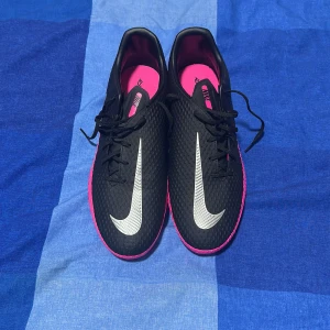 Nike Phantom GT2 svart/rosa fotbollsskor - Nike Phantom GT2 fotbollsskor i svart med starka rosa detaljer och silverfärgad swoosh. Skorna har snörning, platt sula med dobbar och en texturerad ovandel för bättre bollkontroll. Insidan är rosa och märkt med Phantom GT. Perfekt för dig som vill sticka ut på planen.