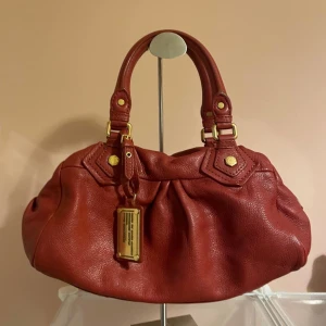 Röd handväska från Marc by Marc Jacobs - Snygg röd handväska i äkta skinn från Marc by Marc Jacobs. Väskan har guldfärgade metalldetaljer, dubbla handtag och ett coolt innerfoder med svartvitt grafiskt mönster. Perfekt storlek för att få plats med allt du behöver och stängs med dragkedja.