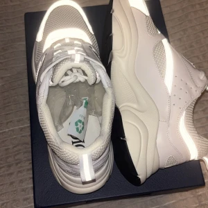 Vita sneakers från Dior - Säljer ett par trendiga vita sneakers från Dior. Skorna har reflekterande detaljer, mesh och skinnpaneler, samt tjock sula i vitt och svart. Snygg design med snörning och diskret Dior-logga på sidan. Perfekta för dig som vill sticka ut med lyxig streetstyle.  PRISET EJ HUGGET I STEN!