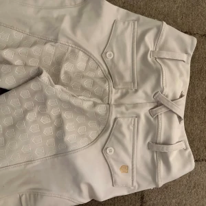 Vita ridbyxor Maya Delorez XXS - Snygga vita ridbyxor från Maya Delorez i storlek XXS. Byxorna har hög midja, mönstrade grip-paneler längs insidan av benen och praktiska fickor med knapp. Materialet är stretchigt syntet för maximal komfort vid ridning.