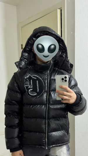 Corvidae - Svart pufferjacka med glansig finish. Jackan har huva, dragkedja framtill och avtagbara ärmar så den kan användas som väst. Perfekt för dig som gillar streetwear och vill ha en jacka som sticker ut. 