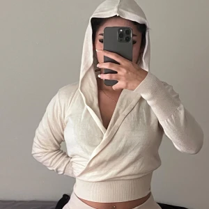 Beige omlott hoodie med huva - Säljer en beige hoodie med omlott-design och breda ribbade muddar. Tröjan har lång ärm och stor huva, perfekt för en chill och stilren look. Materialet känns mjukt och stretchigt, och passformen är normal till lite tight.