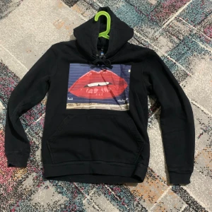 Svart hoodie med tryck från The Cool Elephant - Svart hoodie från The Cool Elephant med stort tryck av röda läppar på bröstet. Tröjan har huva med snörning och en klassisk känguruficka framtill. Perfekt för dig som gillar streetstyle och vill sticka ut med ett coolt motiv.