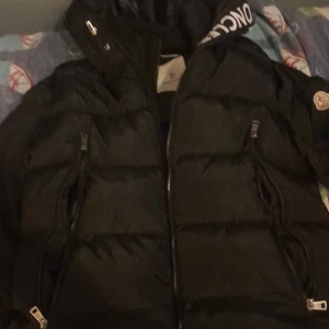 Svart pufferjacka från Moncler - Svart pufferjacka från Moncler med ikonisk logga på ärmen och coolt serietidningsfoder på insidan. Jackan har dragkedjor på fickorna och framtill, samt en hög krage. Perfekt för dig som vill ha en stilren och varm jacka med street vibe.