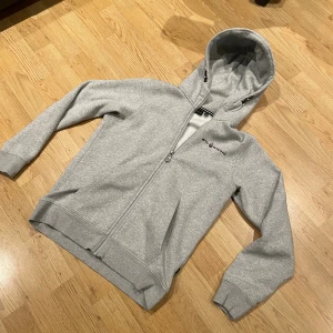 Grå hoodie från Sail Racing - Grå hoodie med dragkedja från Sail Racing. Tröjan har huva med snörning och ett svart tryck på huvan. Logga på bröstet, två fickor fram och ribbade muddar vid ärmslut och nederkant. Perfekt för chill eller häng med kompisar. Den är lätt och stila och passar bra med allt. Det är väldigt bra skick och den är i storlek 160