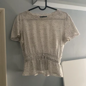 Prickig blus med knyt från Zara - Supersöt vit blus från Zara med svarta prickar, korta ärmar och rund hals. Midjan har en smock med knytband och volangkant nedtill. Är i strl M men passar också någon som har S som jag själv. Säljs då den inte kommer till så mycket användning. 